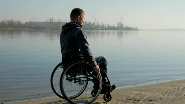 Man-in-wheelchair-rides-in-sand-near-river,-disabled-man-in-wheelchair-near-river