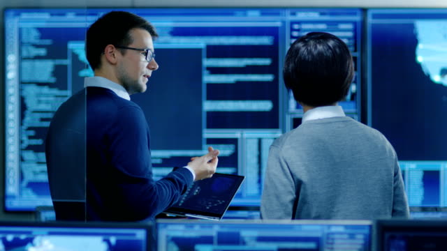 In-the-System-Control-Room-IT-Specialist-and-Project-Engineer-Have-Discussion-while-Holding-Laptop,-they're-surrounded-by-Multiple-Monitors-with-Graphics.-They-Work-in-a-Data-Center-on-Data-Mining,-AI-and-Neural-Networking.