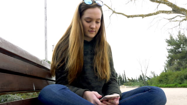 Ein-ernsthaftes-junges-Mädchen-sitzt-auf-dem-Park-Bench-mit-gekreuzten-Beinen-sitzen-und-nutzt-das-Telefon.