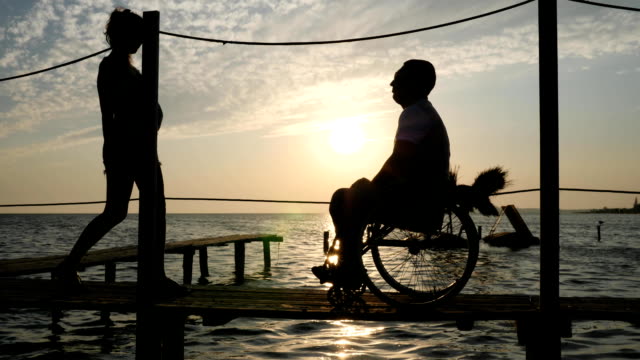 silhouette-of-loving-couple-with-disabled-person-on-seafront-near-shiny-water-in-bright-beams