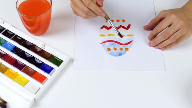 Unrecognizable-woman-paints-a-sketch-with-bright-colors-on-paper-for-an-Easter-card.-On-the-table-is-a-palette-with-paints-and-a-glass-of-water.