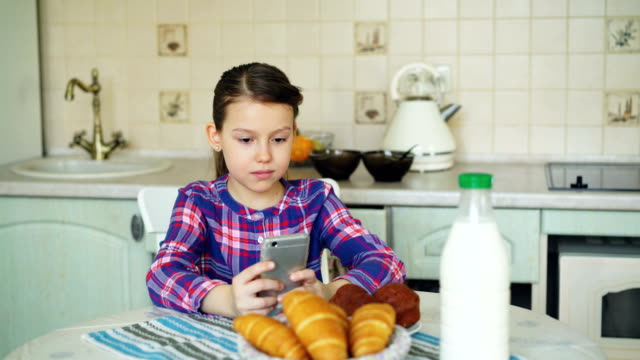 Little-beautiful-girl-playing-smartphone-at-morning-while-sitting-at-table-in-kitchen.-Childhood,-people,-and-technology-concept