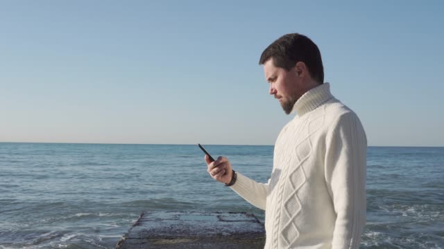 man-holds-a-smartphone-in-his-hands,-the-person-stands-on-a-background-of-waves