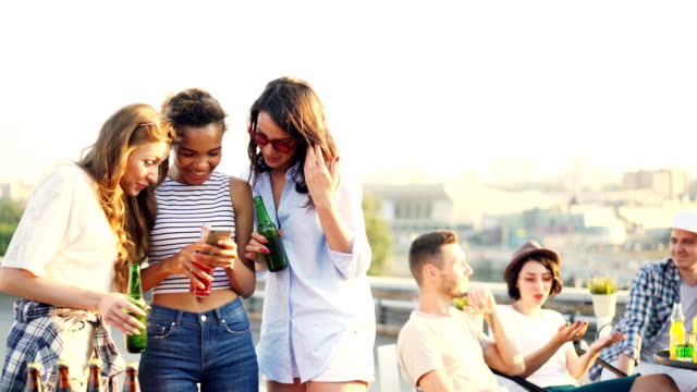 Joyful-female-friends-are-using-smartphone-and-chatting-holding-bottles-during-party-on-roof.-Social-media,-young-people-and-modern-technology-concept.