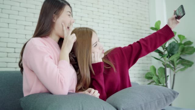 Lesbian-Asian-couple-using-smartphone-selfie-in-living-room-at-home,-sweet-couple-enjoy-funny-moment-while-lying-on-the-sofa-when-relax-at-home.-Lifestyle-couple-relax-at-home-concept.