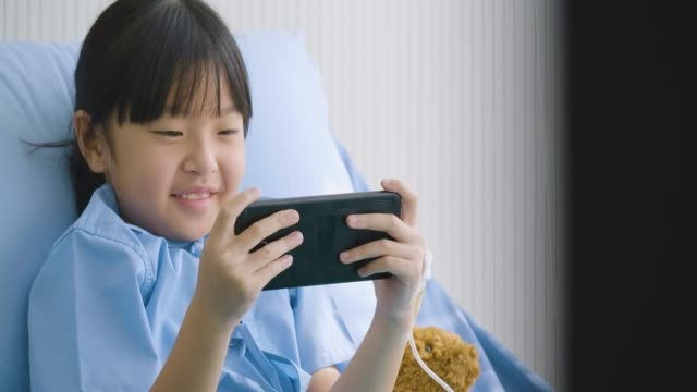 Niedliche-kleine-Mädchen-auf-Bett-im-Krankenhaus,-lustige-Cartoons,-Filme-auf-Smartphone-ansehen.-Krankheit-und-Behandlung.