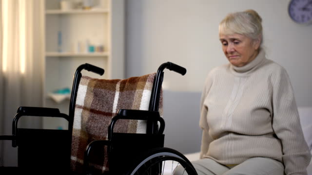 Upset-elderly-woman-sitting-on-sofa-near-wheelchair-in-rehabilitation-center