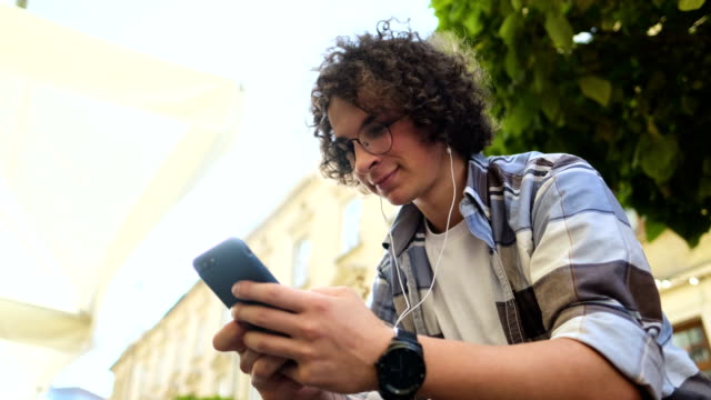 Smiling-man-in-headphones-using-smartphone,-listening-to-music-and-browsing-on-smartphone.-Outdoor.-Close-up.-Blogger-scrolls-through-social-media-on-device,-reading-news-on-app