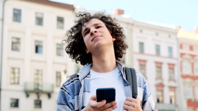 Hübsche-junge-Mann-mit-seinem-Smartphone-scrollt-durch-Social-Media-Feeds-auf-dem-Gerät,-lächelt-von-Nachrichten-über-Anwendungen-und-schaut-auf-die-Kamera-auf-der-Straße,-im-Freien