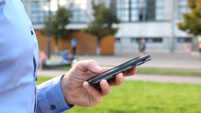 Close-up-hand-of-young-businessman-holding-and-touching-smartphone.-Unrecognizable-man-standing-on-urban-street-and-browsing-phone.-Guy-using-gadget-for-work.-Slow-motion-Side-view