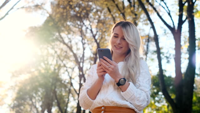 Portrait-of-girl-in-white-sweater-writing-message-on-her-smartphone-outdoors.-Woman-using-digital-gadget,-scrolls-through-social-media,-reading-news-on-app,-at-sunset.-Cute-girl-in-city-park
