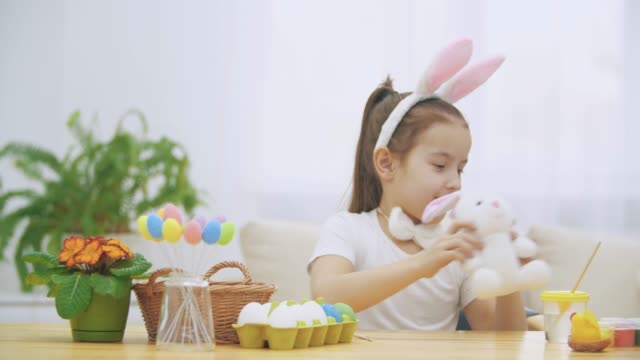 Brisk,-cute-girl-is-having-fun-painting-with-Easter-bunny,-which-has-the-same-ears,-as-she-has.-Girl-is-setting-back-white-bunny.-Girl-with-a-beauty-spot-at-her-face-and-is-smiling-widely,-sitting-at-the-wooden-table-with-Easter-decorations.