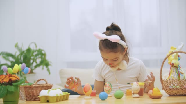 Cute-girl-is-having-fun-painting.-Adorable-girl-is-colorizing-an-Easter-egg-and-her-hands-with-a-help-of-paint-brush-in-her-mouth.-Girl-with-beauty-spots-at-her-face-and-is-looking-gently-with-cat's-whisker,-sitting-at-the-wooden-table-with-Easter-decorat