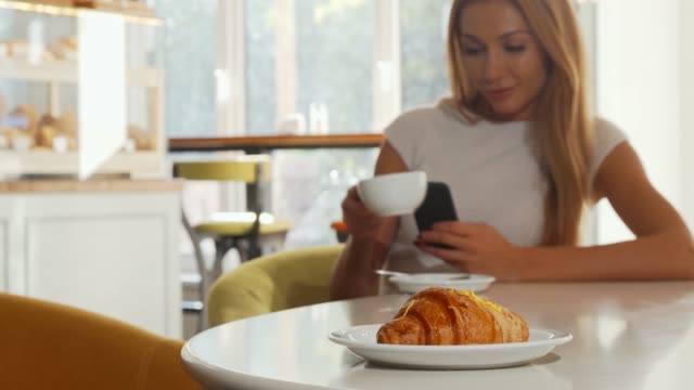 Enfoque-selectivo-en-croissant-sabroso,-mujer-usando-teléfono-inteligente-en-la-cafetería