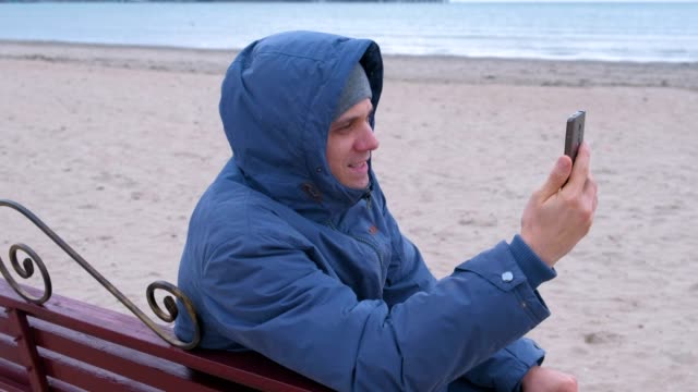 Man-blogger-in-a-blue-down-jacket-sitting-on-a-bench-on-the-sand-beach-and-talking-a-video-chat-on-mobile-phone.