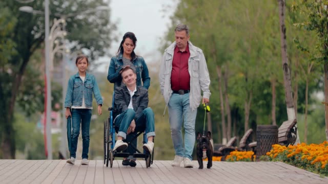 Cuidar-a-la-familia-con-un-adolescente-discapacitado-camina-por-el-parque-en-una-tarde-de-otoño.-Joven-en-silla-de-ruedas-pasa-el-día-en-el-parque-con-un-perro-y-los-padres.