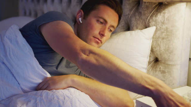 Hombre-en-la-cama-poniendo-auriculares-inalámbricos-conectados-al-teléfono-móvil-antes-de-ir-a-dormir