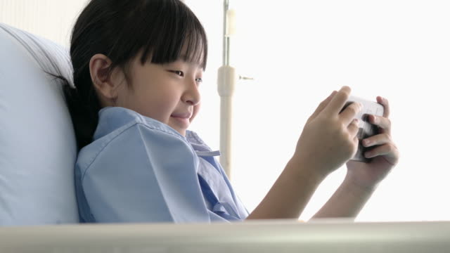 Asiatische-kleine-Mädchen-lag-im-Krankenhausbett-und-Emotion-glücklich-spielen-Videospiel-online-auf-dem-Smartphone.-Das-kranke-Mädchen-ist-patientim-im-Krankenhaus-und-in-moderner-medizinischer-Versorgung.