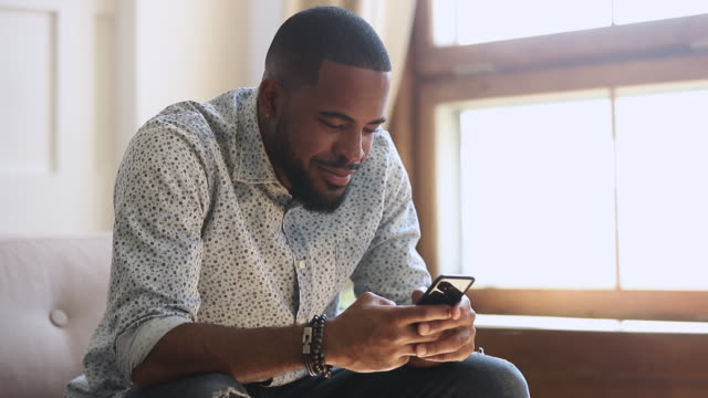 Sonriendo-hombre-afroamericano-sosteniendo-mensajes-de-texto-de-teléfono-inteligente-en-casa