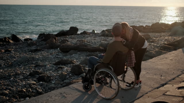 Gesundes-Mädchen-umarmt-ihren-kranken-Freund-im-Rollstuhl-auf-See-Kai