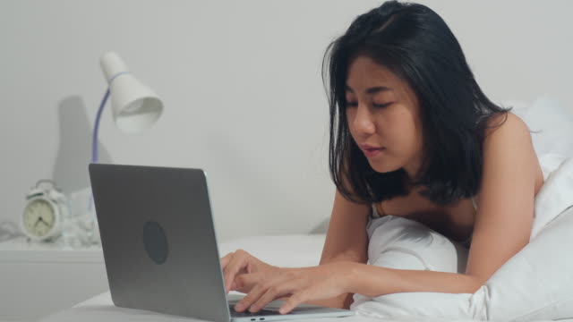 Junge-asiatische-Frau-mit-Laptop-Überprüfen-Social-Media-Gefühl-glücklich-lächeln,-während-auf-dem-Bett-liegen-duponieren,-nachdem-sie-morgens-im-Haus-aufwachen,-attraktive-thai-Mädchen-lächelnd-entspannen-im-Schlafzimmer-zu-Hause-Konzept.