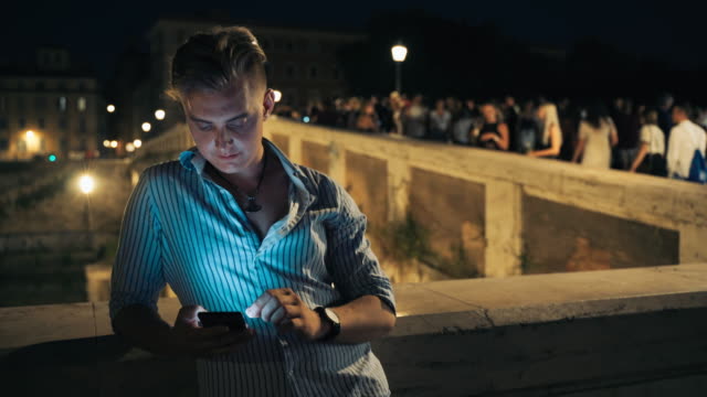 Young-handsome-man-using-his-smartphone-at-night.-Man-staying-in-the-city-centre-and-enjoying-night-life.-Tourist-male-browsing-internet-on-background-of-crowded-street-at-night.-Illuminated-night-street-full-of-walking-people