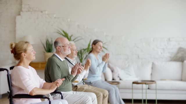 Side-view-of-group-of-four-senior-patients-including-disabled-ones-applauding-to-speaker-during-entertainment-time-in-nursing-home,-focus-on-smiling-bearded-man-in-eyeglasses