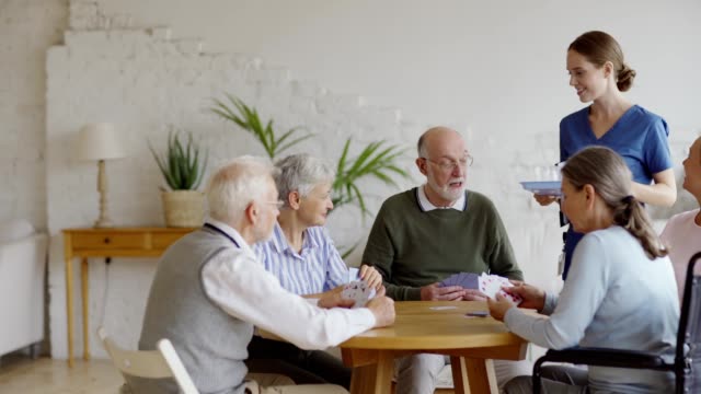 Young-nurse-bringing-vitamin-pills-and-glass-of-water-for-senior-man-playing-cards-with-aged-friends-sitting-at-table-in-common-room-of-nursing-home,-tracking-shot