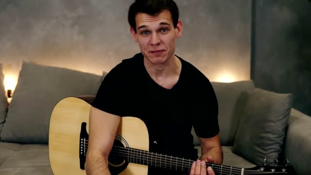 Portrait-of-short-haired-caucasian-man-in-black-T-shirt-making-video-blog-about-musical-instruments-or-recording-the-song-for-his-followers-while-sitting-on-a-couch-at-home.-Looking-to-the-camera