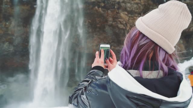 schöne-junge-Frau-in-der-Nähe-eines-Wasserfalls-in-Island,-macht-Fotos-am-Telefon,-freut-sich-und-macht-ein-Selfie