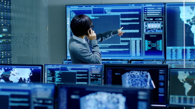 In-the-System-Control-Room-IT-Administrator-Talks-on-the-Phone-with-His-Superiors.-He's-in-a-High-Tech-Facility-That-Works-on-the-Surveillance,-Neural-Networks,-Data-Mining,-AI-Projects.