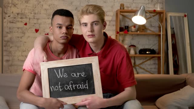 A-sad-international-gay-couple-is-sitting-on-the-couch-and-holding-a-sign.-We-are-not-afraid.-Look-at-the-camera.-Home-comfort-on-the-background.-60-fps
