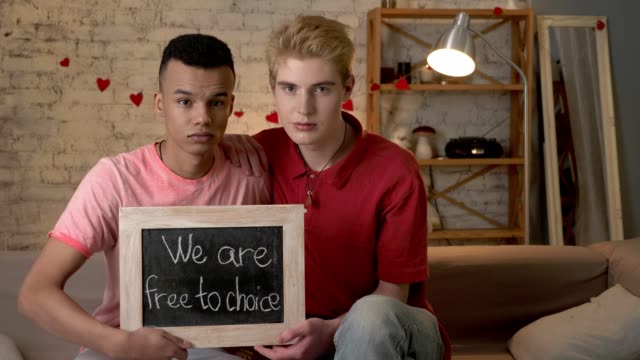 A-sad-international-gay-couple-is-sitting-on-the-couch-and-holding-a-sign.-We-are-free-to-choise.-Home-comfort-on-the-background.-60-fps