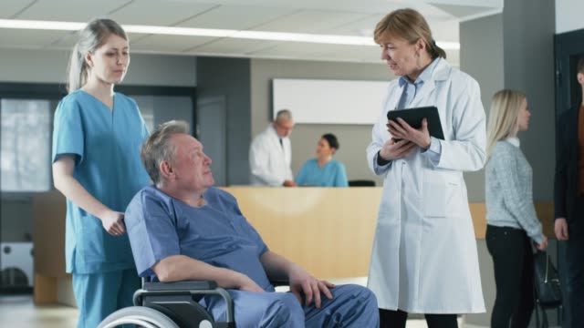 In-der-Klinik-Lobby,-Krankenschwester-schiebt-älteren-Patienten-im-Rollstuhl,-zwar-Arzt-spricht-mit-ihnen-mittels-Tablet-Computer.-Sauberes,-neues-Krankenhaus-mit-medizinischen-Fachpersonal.-Slow-Motion.