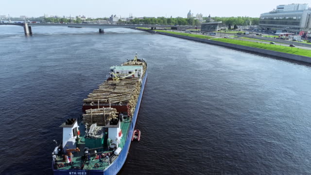 Barge-with-wood-is-pulling-up-big-blue-river-through-city-with-bridge