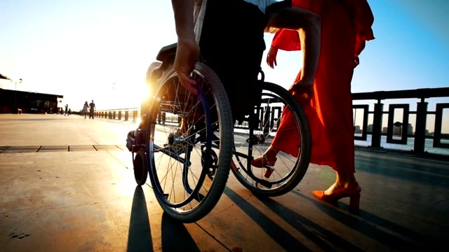 Kerl-mit-Kranken-Beinen-am-Rollstuhl-zu-Fuß-mit-Freundin-auf-der-Uferpromenade-In-den-Sommerabend