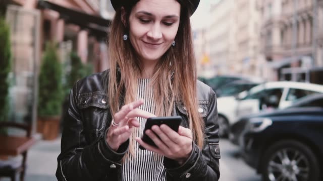 Lächelnd,-glücklich-kaukasischen-Bloggerin-in-stilvollen-Hut-zu-Fuß-entlang-einer-Straße-mit-Smartphone-app-Slow-Motion-einkaufen