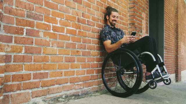 Young-happy-disabled-man-texting-on-phone-in-wheelchair-leaning-against-brick-wall