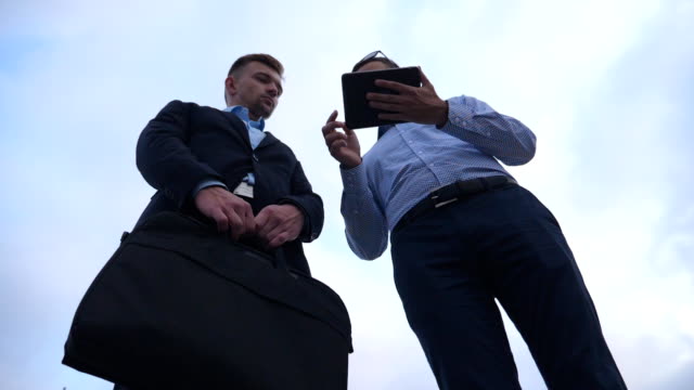Two-young-businessmen-talking-and-using-tablet-pc-in-city.-Handsome-coworkers-discussing-and-working-on-digital-tablet-with-sky-at-background.-Colleagues-applying-mobile-technology.-Low-angle-view