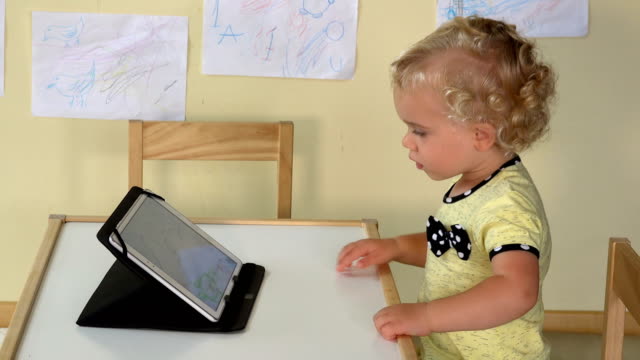 niedliche-Baby-Mädchen-mit-Tablet-Computer-sitzen-in-der-Nähe-von-einem-kleinen-Tisch.