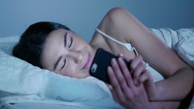 Eine-Frau-in-ihrem-Bett-und-nutzt-ihr-Telefon,-um-Nachrichten-aus-der-Ferne-zu-verschicken,-Videoanrufe-zu-verschicken-und-futuristische-Grafiken-zu-erscheinen.