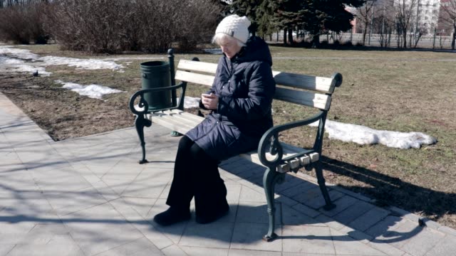 A-senior-elderly-woman-sitting-on-a-bench-in-park-outdoors-and-using-tapping-smartphone--she-is-useing-smartphone-to-connect-with-people-on-social,-texting-messages-ang-searching-internet