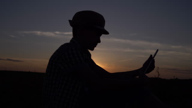 Silhouette-eines-Jungen-nutzt-bei-Sonnenuntergang-auf-dem-Feld,-liest-etwas-auf-dem-Tablet,-im-Freien