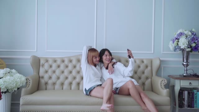 Happy-couple-friend-sitting-on-sofa-hug-and-using-phone-taking-selfie-together-at-home.-Friend-and-lesbian-concept.