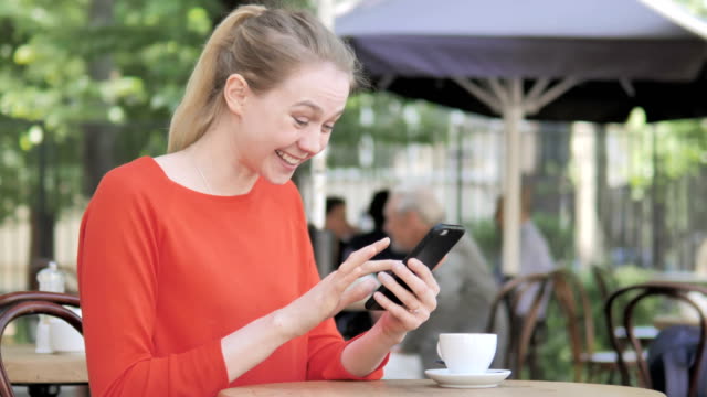 Mujer-joven-aclamación-éxito-en-Smartphone-sentado-en-Cafe-Terrace