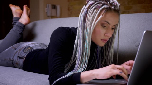 Closeup-shoot-of-young-attractive-caucasian-hipster-female-with-dreadlocks-using-the-laptop-lying-on-the-couch-indoors-in-a-cozy-apartment