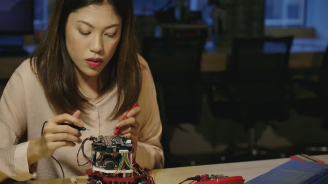 Las-jóvenes-ingenieras-de-desarrollo-electrónico-asiático-trabajan-con-el-robot,-midiendo-la-señal-en-los-circuitos-eléctricos-del-prototipo-de-robótica-en-el-taller.-Personas-con-concepto-de-tecnología-o-innovación.