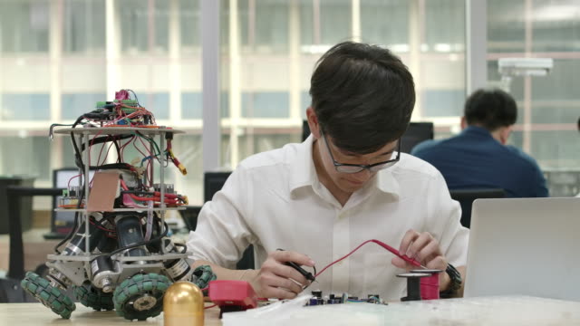 Young-asian-electronics-development-engineers-works-with-robot,-measuring-the-signal-in-the-electrical-circuits-of-robotics-prototype-in-workshop.-People-with-technology-or-innovation-concept.
