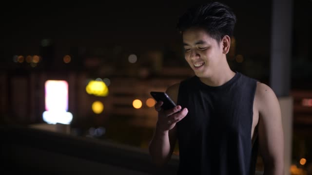 Portrait-of-young-happy-Asian-man-using-phone-outdoors-at-night