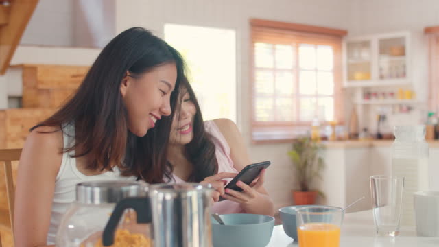Asiatische-Lesbische-Paar-mit-Handy-Check-News,-während-frühstücken-in-der-Küche-zu-Hause.